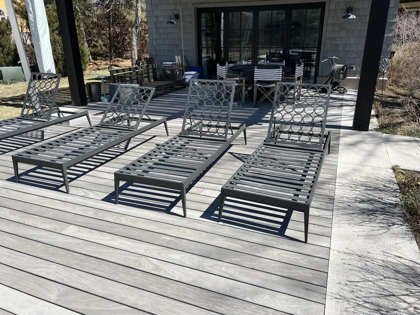 Grey deck boards meeting a stone-paved walk beside a shingled house, with loungers in the sun